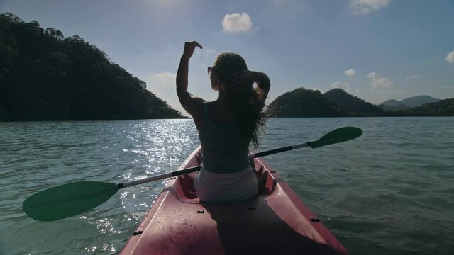 Silhouette of woman tourist raising hands while sailing in canoe along sea bay water to distant hilly island backside view.