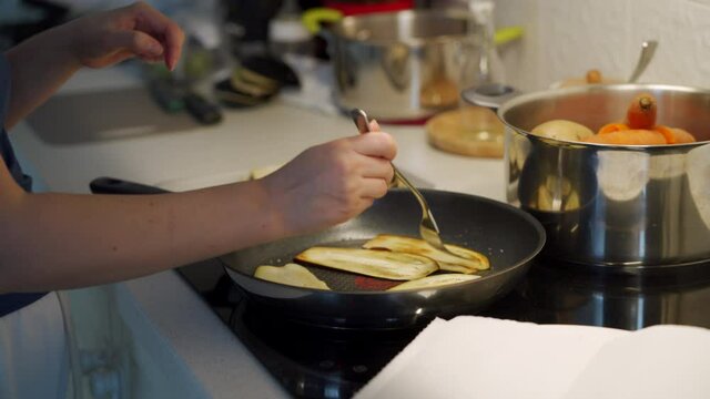 Cooking on electric stove top in the kitchen, woman fries eggplant wedges in frying pan on induction cooktop closeup. High quality 4k footage