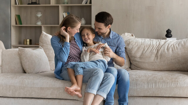 Loving caring happy young couple parents tickling adorable little preteen kid daughter, having fun sitting on cozy couch together in modern living room, enjoying carefree playtime weekend activity.