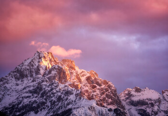 Beautiful mountain peaks in snow and violet sky with pink clouds in winter at sunset. Colorful landscape with high snowy rocks, cloudy purple sky in cold evening. Alpine mountains. Nature background