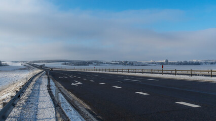 Fototapeta premium Country road through winter rural scene. Black clean asphalt. Winter road going through snow-covered fields.