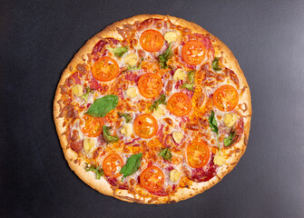 Freshly baked pizza with several types of sausage, ham, cherry tomatoes, mushrooms, basil leaves and lettuce on a black background.