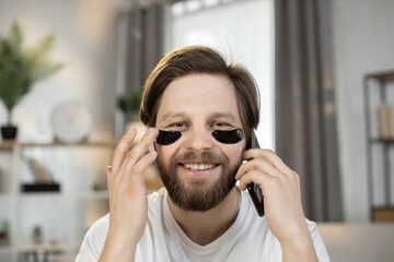 Close up of modern busy young bearded Caucasian man, with black hydrogel patches under eyes, sitting at home and talking on the phone with friend