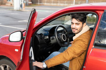 Portrait of hispanic or eastern ethnicity smiling man in jacket opens door of red car on parking slot on autumn, winter or spring day. Travel, exam, lesson, learning, taxi driver. View from outside.