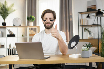 Front view of young handsome Caucasian bearded man, with black collagen eye patches, sitting at the table at home showing thumb up and working on laptop remotely. Online work and beauty procedures