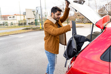 Young latin or arab eastern indian pakistani man taking out or putting backpack in trunk of red...