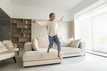 Overjoyed energetic happy little child girl jumping from comfortable couch on floor, having fun...