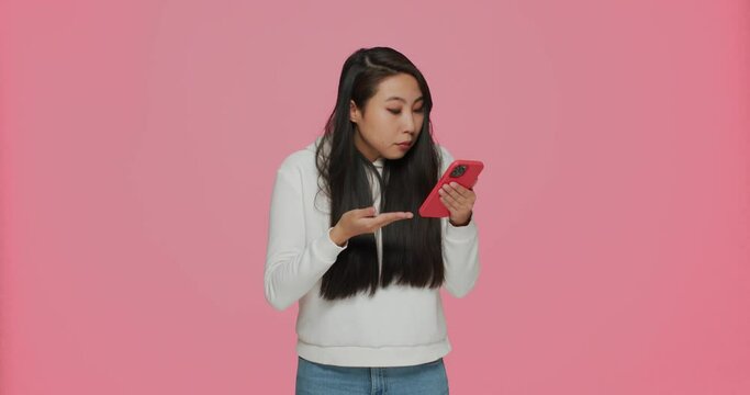 Puzzled confused young asian girl reading spam message holding phone, shrug shoulders on pink studio background
