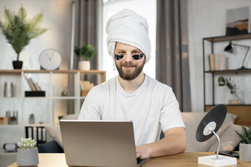 Front view of good-looking smiling young bearded man with hydrogel black patches under eyes and towel on his head, sitting at the table at home and typing on laptop