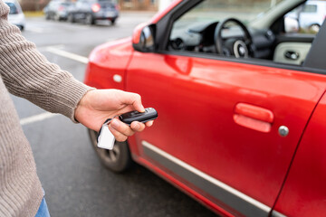 Male hand of young man holding electronic remote key pushing button near red rental car to open or...