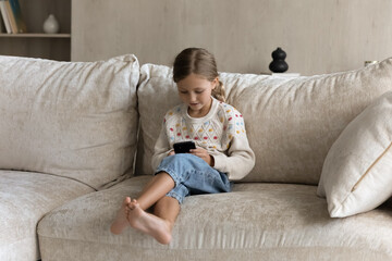 Fototapeta Full length joyful small Z generation cute kid girl using smartphone applications, resting on cozy couch, enjoying web surfing information, playing online games or watching funny video alone at home. obraz