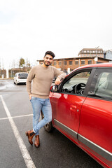 Fototapeta na wymiar Young latin or arab eastern indian pakistani carefree man in stylish clothes standing near red small car on parking slot of an urban city background on rainy midseason day. Travel, rental, car sharing