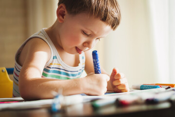 The boy is sitting at the table and diligently draws something with felt-tip pens. The child is trying so hard that he stuck out his tongue. - obrazy, fototapety, plakaty