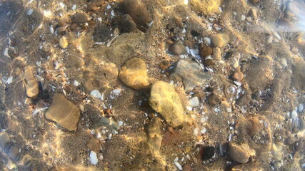 Background yellow sandy bottom with stones rocks, seashells close-up. Movement lines stripes of sun glare along seabed. Natural sunny backdrop. Motion on bottom solar reflection ripples on water. Wave