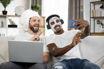 Men skin care at home. Two young multiethnic men making facial hygiene procedures, using eye patches and facial mask making selfie photo on smartphone. Gay couple at home