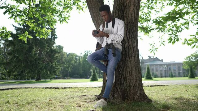 Wide shot happy talented African American photographer standing at tree leaning on trunk watching photos on camera smiling as squirrel running at background. Joyful confident man outdoors on sunny day