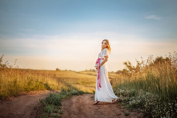 A pregnant woman in a white dress stands barefoot on a path in a field on a sunny summer day.