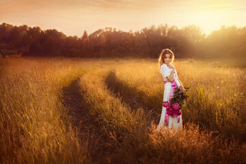 A pregnant woman in a white dress with a bouquet of peonies in a field at sunset.