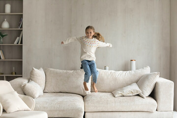 Full length excited cheerful laughing barefoot small 7s kid girl jumping on sofa, dancing to music, enjoying leisure domestic activity, having fun entertaining alone in living room, playtime concept.