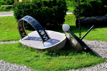 A close up on a sun dial and sun clock made out of metal and stone, located next to an old abandoned anchor of a big ship located in the middle of a dense public park next to a gravel path or road - Powered by Adobe