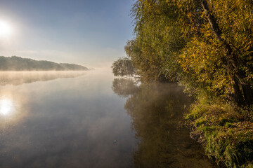 Autumn landscape in the early morning on the river. Misty water surface. Yellow leaves on trees and bushes are illuminated by the rays of the rising sun. Dawn on a cold autumn morning.