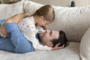 Smiling caring young father cuddling cute little kid daughter, enjoying funny conversation, talking chatting having fun lying together on comfortable sofa at home, sincere loving family relations.