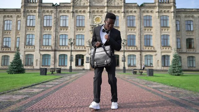 Wide shot portrait of joyful graduated African American student in formal suit with attache bag walking dancing on university yard outdoors. Dolly shot happy smart man rejoicing university graduation