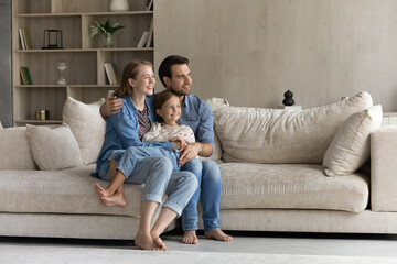 Happy loving young couple parents and little adorable kid daughter resting on comfortable sofa, looking in distance, visualizing or planning future, remembering good moments at home, copy space.