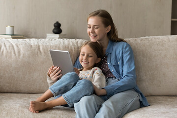 Happy bonding young mother and laughing cute kid daughter looking at digital computer tablet screen, shopping in internet store, playing online games or watching funny cartoons, modern tech addiction.