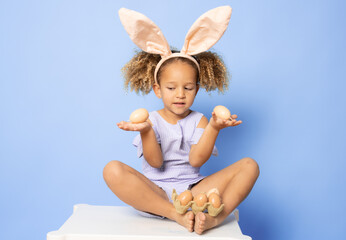 Portrait of cute little girl with Bunny ears and Easter eggs sitting on a table over purple...