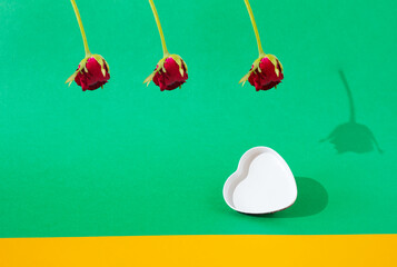 Three red roses with a shadow hanging above a white heart on a green and orange background. Valentine's Day. Minimalist concept of love. Copy space.
