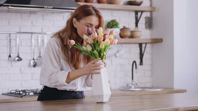 cheerful adult woman with long red hair holding vase with flowers cozy home in spring kitchen interior