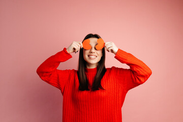 Cute girl is smiling and covering her face with paper hearts. The girl is standing isolated against the pink background. Concept of the St. Valentine's Day