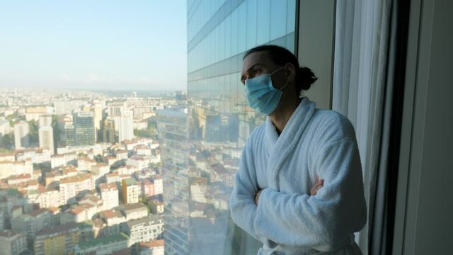 Tired of being quarantined, man stands in his bathrobe and face mask at window and looks out over inaccessible city. Traveler on mandatory quarantine after arrival in another country