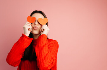 Cute girl is sending a kiss and covering her face with paper hearts. The girl is standing isolated against the pink background. Concept of the St. Valentine's Day