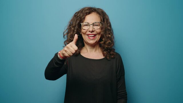 Middle age woman smiling confident doing ok gesture with thumbs up over blue background