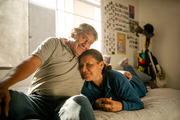 Retrato frontal de pareja de esposos latinos abrazados mirando a cámara con afecto  sobre su cama 