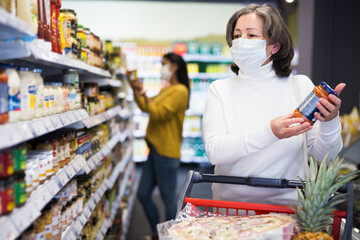 Woman in protective mask with shopping cart choosing groceries in supermarket