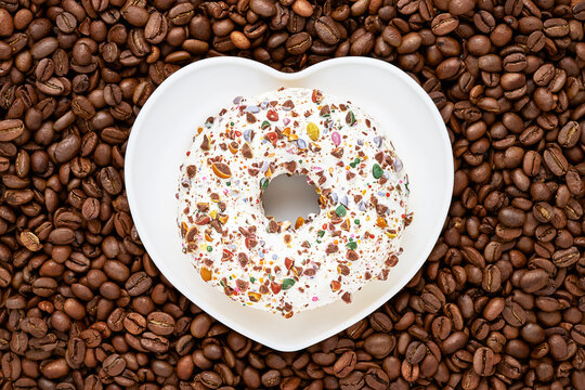 Donut in white glaze with candy sprinkles lays on a heart shape plate over coffee beans background