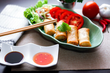 Fried spring rolls, vegetables and tomatoes placed in a green leaf shape plate on a black wooden table and dipping sauce.