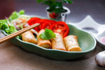 Fried spring rolls, vegetables and tomatoes placed in a green leaf shape plate on a black wooden table and dipping sauce.
