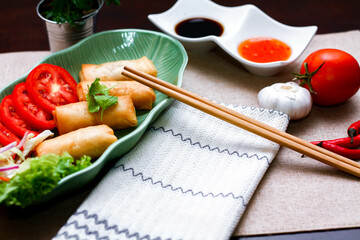 Fried spring rolls, vegetables and tomatoes placed in a green leaf shape plate on a black wooden table and dipping sauce.