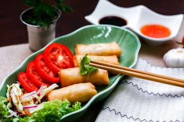 Fried spring rolls, vegetables and tomatoes placed in a green leaf shape plate on a black wooden table and dipping sauce.