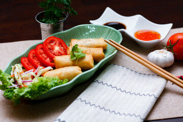 Fried spring rolls, vegetables and tomatoes placed in a green leaf shape plate on a black wooden table and dipping sauce.