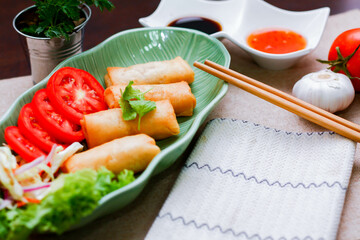 Fried spring rolls, vegetables and tomatoes placed in a green leaf shape plate on a black wooden table and dipping sauce.