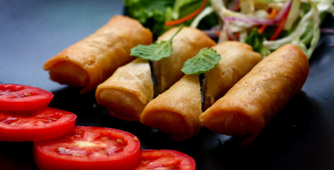 Fried spring rolls with vegetables and tomatoes placed in a black plate on a black wooden table and dipping sauce.