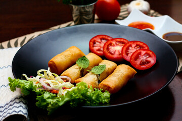 Fried spring rolls with vegetables and tomatoes placed in a black plate on a black wooden table and dipping sauce.