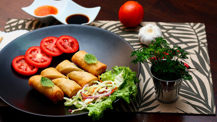 Fried spring rolls with vegetables and tomatoes placed in a black plate on a black wooden table and dipping sauce.