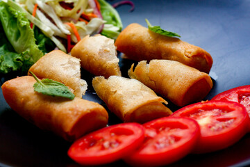Fried spring rolls with vegetables and tomatoes placed in a black plate on a black wooden table and dipping sauce.