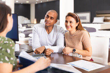 Young Latin american couple visiting furniture store in search of comfortable mattress, talking to saleswoman